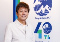 FMヨコハマ開局40周年の記念ロゴパネルの横で、白いシャツを着て微笑むDJの栗原治久さんのポートレート。