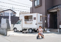 八潮市の米粉のぱん屋「てとてとて」の移動販売車（キッチンカー）の外観。白い車体と、手前に三輪車に乗った子どもがいる。