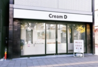 「Cream D Kawasaki」の店舗外観。白地に黒のシンプルなロゴ看板が掲げられ、大きなガラス張りの入り口の横にはメニューが記された立て看板が置かれている。