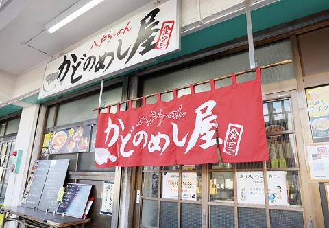 足立市場内にある「かどのめし屋 海鮮食堂」の店舗外観。赤地に白文字で店名が書かれた大きな暖簾がかかり、上部には「八戸らーめん」と記された看板がある。