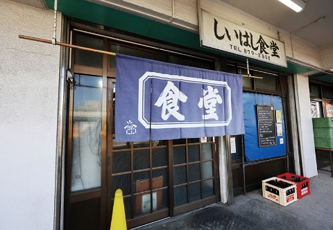 足立市場内にある「しいはし食堂」の店舗外観。入り口には「食堂」と大きく書かれた紺色の暖簾がかかり、上部には店名と電話番号が記された看板がある。
