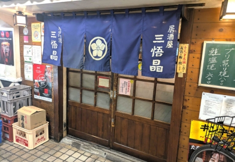 さいたま市大宮にある居酒屋「三悟晶（さんごしょう）」の入り口。紺色の暖簾に白抜きで店名と家紋がデザインされている。